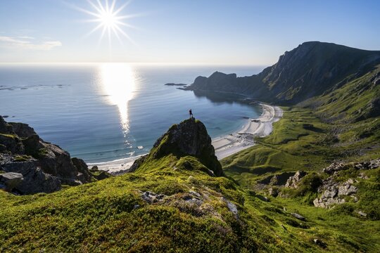 Climber on a rock, view of picturesque coastal landscape with sun star, steep coast with peak M&aring;tind or M&aring;tinden, H&oslash;yvika beach and sea, hike to the summit of M&aring;tind, And&oslash;ya island, Vester&aring;len, Nordland, Norway