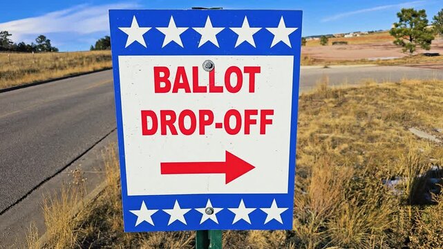 Election ballot drop-off sign fills the frame beside a paved road, featuring bold red lettering, patriotic stars, and an arrow directing voters to the official collection site nearby.