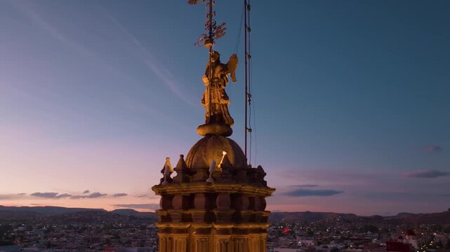 A striking close-up aerial shot capturing the ornate angel statue atop a Durango cathedral silhouetted against a radiant golden hour sky.