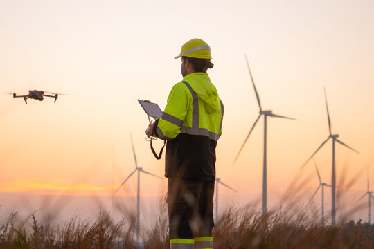 Engineers male flying drone surveying and checking wind turbines from the high angle view of the field during beautiful sunset. using drone technology for work.
