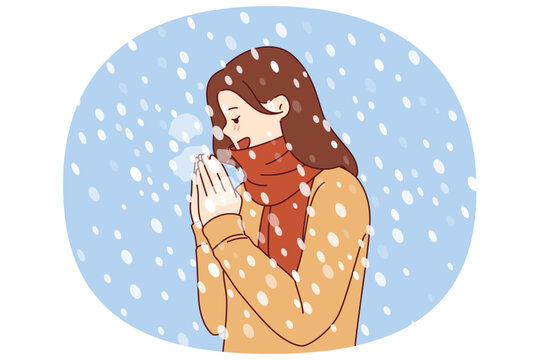 Woman enjoys winter weather, stands under falling snow flakes in sweater and scarf. Positive girl rejoices at onset of winter and approach of christmas holidays, blowing on hands to keep warm