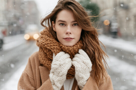 Woman stands snowy street, wrapped in warm scarf and gloves. Soft snowflakes drift around her as wind plays with her hair. She gazes calmly at camera, serene expression in wintry setting