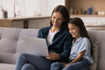Fototapeta premium Happy young mother and cute school-age daughter, making purchases through internet store, enjoy on-line shopping resting on sofa in living room, laughing, watching comedy movie or cartoons on laptop