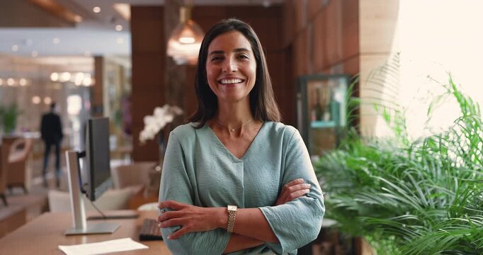 Head shot portrait of smiling Brazilian woman, hotel owner, executive chief standing confidently in modern hotel registration desk with welcoming expression, radiates professionalism and hospitality