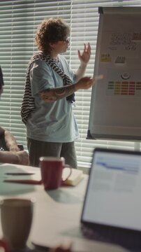 Young female manager with tattoos and yellow glasses pointing to graphics on flipchart and explaining presentation while leading team meeting in office. Vertical shot
