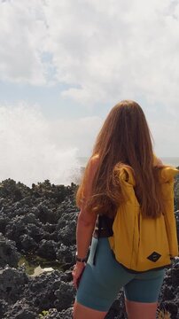 Vertical video. Female Explorer Observing Powerful Ocean Spray Erupting From Sharp Coastal Rocks in Slow Motion