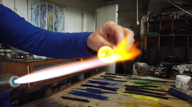 Close up of artist hands creating glass pendant with lampworking technique. Shaping glass with flame making jewelry using gas burner