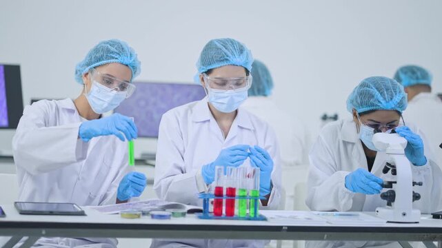 Scientists in masks and protective caps working at bench, one holding green test tube, one writing notes and one using microscope, science team.