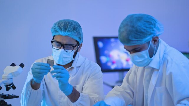 Two researchers in protective caps and masks examining tiny specimen together, microscope in foreground, focused laboratory work.