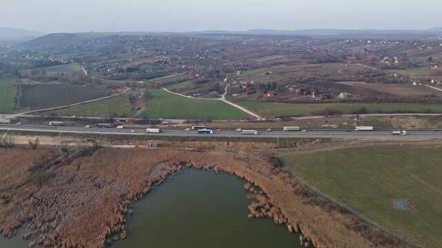 Bicske, Hungary, M1 motorway traffic with cars and trucks near a fishing lake, farmland and residential areas in the Hungarian countryside, viewed by drone.