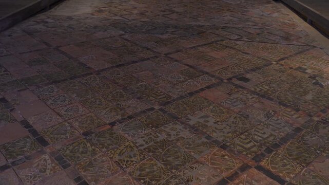 Slow cinematic pan across medieval encaustic floor tiles inside Cleeve Abbey, showcasing intricate geometric patterns and preserved craftsmanship, surrounded by textured stone walls.
