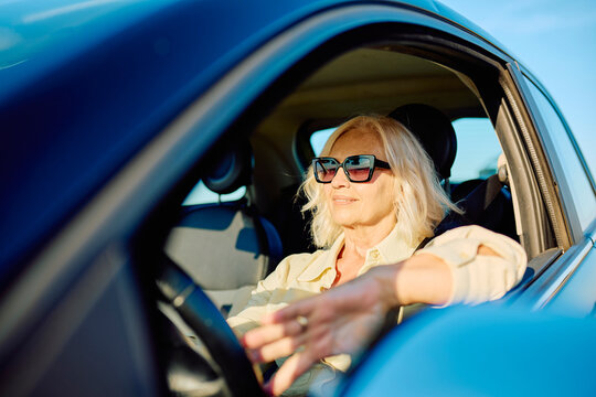 Blond senior woman wearing sunglasses driving a car with an open window, feeling the breeze and enjoying freedom on a sunny day