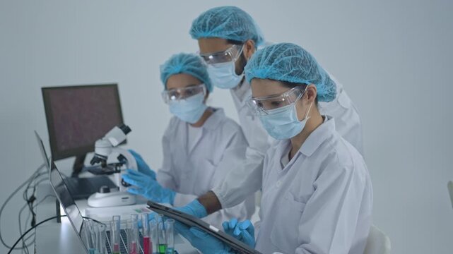 Scientists in masks and protective caps analyzing samples at desk with microscope and computers, one holding sample tube, lab teamwork.