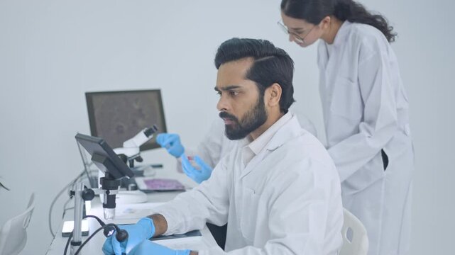 Male scientists using touchscreen of digital microscope at desk while female colleague gestures behind, advanced lab equipment concept.