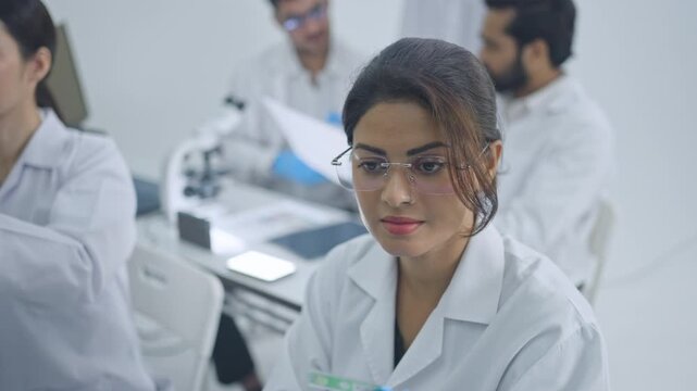 Young woman in eyeglasses and lab coat looking down at work while research colleagues discuss documents in background, bright laboratory.