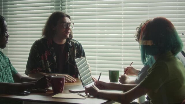 Gen Z creative businessman in casual floral shirt and glasses explaining business plan to diverse team of colleagues during meeting in office