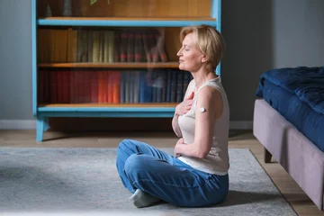 Fotobehang Lotusbloem Diabetes and mental wellbeing. Calm serene mature woman with glucose sensor on arm sitting in lotus pose meditating doing breathing exercises at home  © memento_jpeg