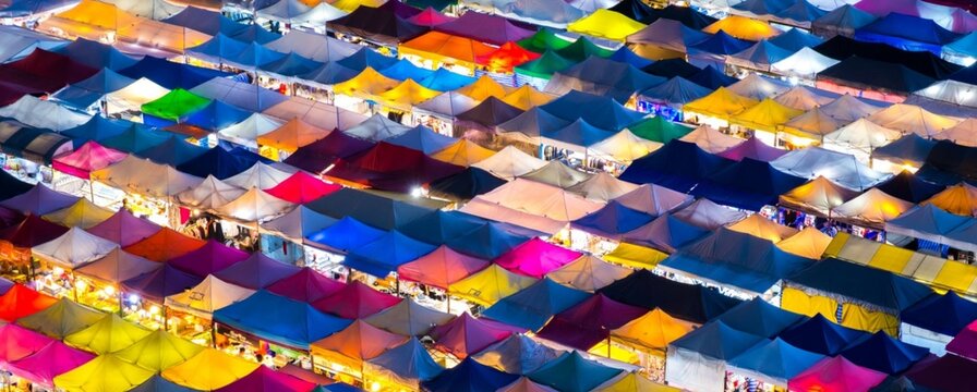 Vibrant tents fill the market square below. Colors glow under warm evening light. Crowds move between stalls and booths. Festive energy pulses through the scene. Ideal for event, travel