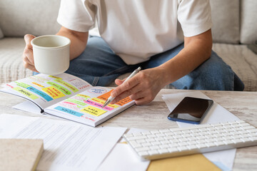 Person holding a pen and a mug, organizing appointments, expenses, and deadlines in a calendar planner book with colorful sticky notes, managing personal life and finances © Nanci