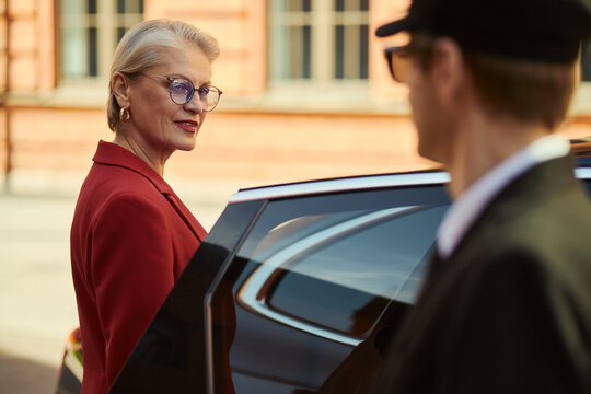 Middle aged Caucasian businesswoman entering car while driver assisting curbside, representing executive travel and leadership. Ideal for corporate mobility, VIP service, business transport