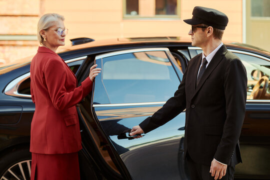 Senior Caucasian businesswoman entering chauffeured car while driver opening door, representing executive travel and premium service. Ideal for luxury transport marketing and VIP mobility