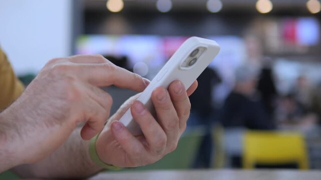 Close-up of hands using smartphone indoors. Fingers tap screen actively. Background shows blurred public space. Person focused on device. Suitable for tech, communication, or mobile usage themes