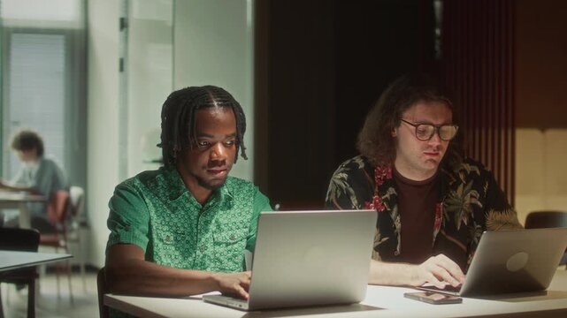 Young African American man and his Caucasian colleague sitting side by side at desk, working on their laptops in modern open space office