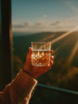 Golden sunlight pours through mountains, illuminating glass filled with refreshing drink in hand