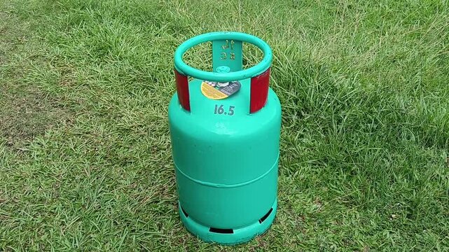 A green 16.5 kg LPG gas cylinder placed upright on a grassy field outdoors. The steel liquefied petroleum gas cylinder features a protective ring around the valve . in malaysia