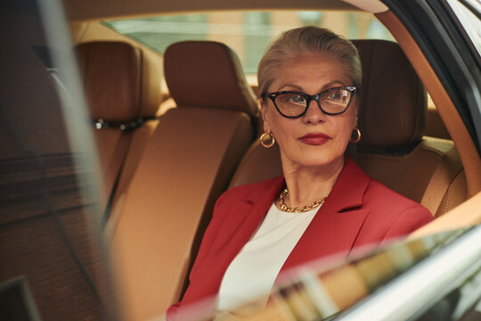 Senior Caucasian businesswoman sitting in car, looking through window while commuting to meeting, conveying executive confidence and mobility. Ideal for leadership or luxury service marketing