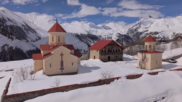 Amaglebis Church complex in Gudauri surrounded by snow-covered terrain, with dramatic Caucasus mountain peaks rising in the background.