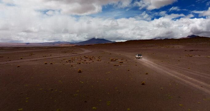 Off road vehicle crossing Siloli Desert in Bolivia beneath dramatic clouds, drone dynamic rearview tracking across empty plains as dust rises