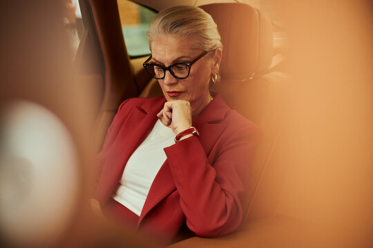 Middle aged Caucasian businesswoman sitting in car, reviewing smartphone with focused expression, representing executive mobility and decision making. Corporate travel marketing