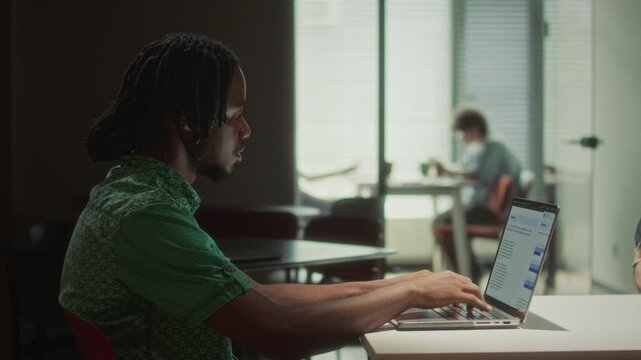 Focused young Black businessman with dreadlocks typing on laptop at desk in dark office late in evening. Side view