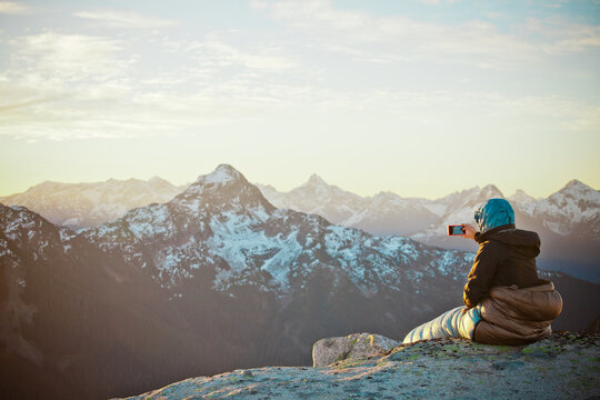 Hiker photographs sunrise from sleeping bag on mountain summit