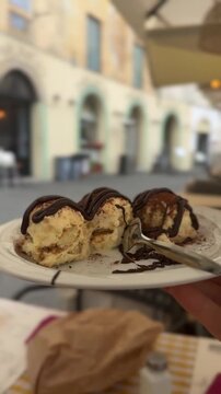 Hand holds a plate with a traditional Italian cream dessert topped with chocolate while seated at an outdoor cafe . The dessert is presented to the camera, highlighting its texture and ingredients bef