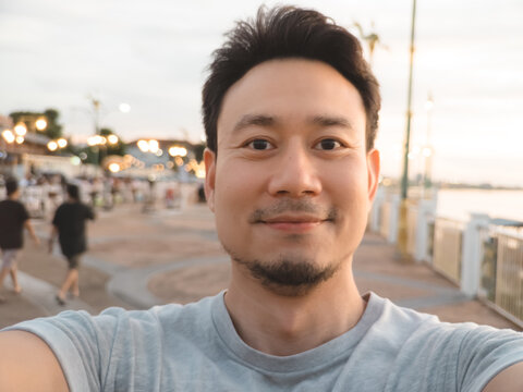 Happy Asian man taking a selfie outdoors during a relaxed evening stroll by the waterfront.