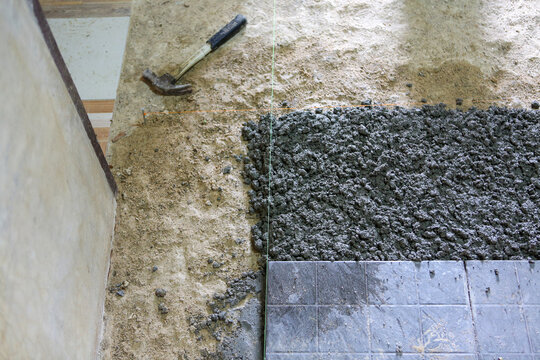 Wet cement floor being laid next to ceramic tile with hammer on rough subfloor, renovation scene showing texture and work in progress, gritty and practical atmosphere