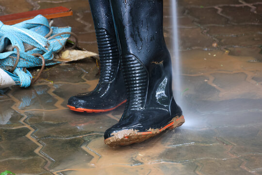 Black work boots being sprayed clean with high pressure stream on wet muddy pavement, showing water splash and dirt removal