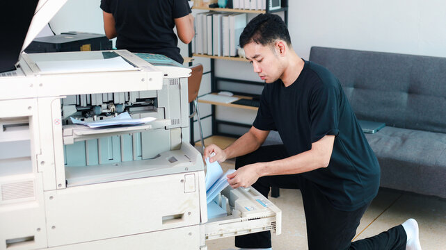 Office technician inspecting an open commercial photocopier while holding printed documents, representing printer repair, maintenance, troubleshooting,