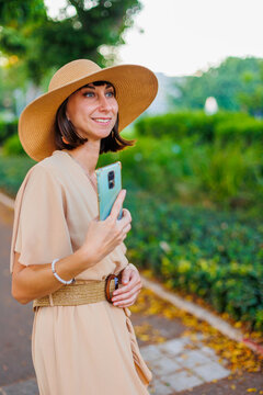 A young woman in a dress and summer hat takes a photo.