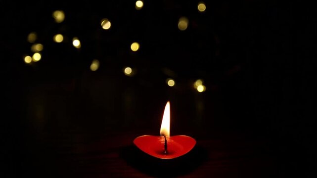 Small red heart-shaped candle is lit on dark wooden surface, its flame ignites and grows steadily brighter against glowing golden bokeh lights in a cozy evening setting.