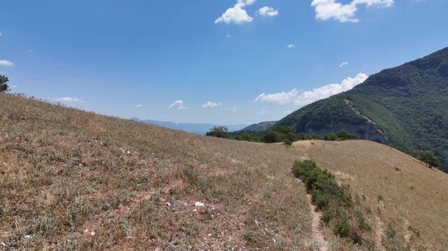 Pendio con erba secca e arbusti isolati che si apre verso montagne verdi e vallate naturali, sotto un cielo limpido. Paesaggio ampio, luminoso e incontaminato