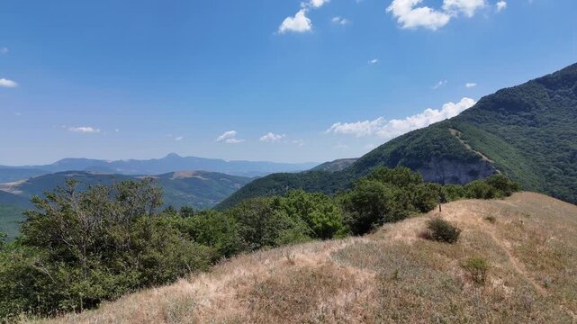 Pendio con erba secca e arbusti sparsi che si estende verso colline verdi e montagne sovrapposte, sotto un cielo azzurro con poche nuvole. Paesaggio naturale ampio, luminoso e incontaminato