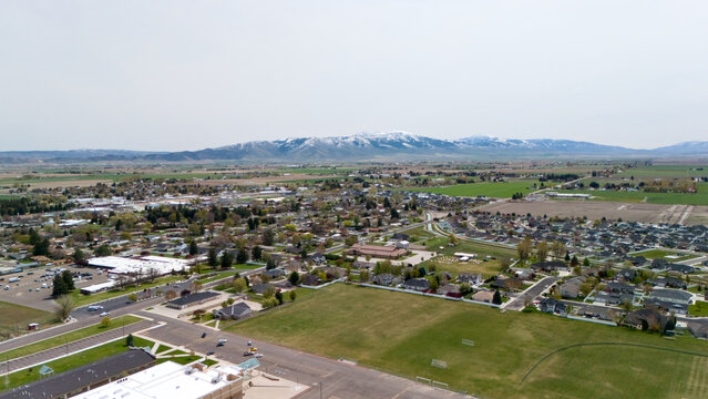 Aerial view of an open green field bordering a growing residential subdivision near mountains.