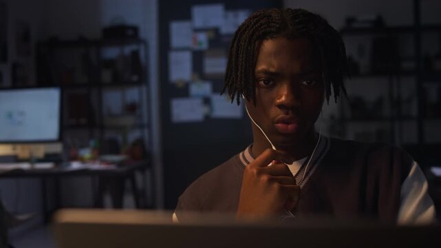 Young adult African American man wearing earphones looking intently at computer screen during online call while working late in office. Footage for corporate training or cybersecurity campaigns