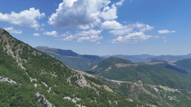 Catena montuosa verde con pendii ricoperti di foresta, crinali sovrapposti e una strada che attraversa il paesaggio, sotto un cielo azzurro con nuvole bianche