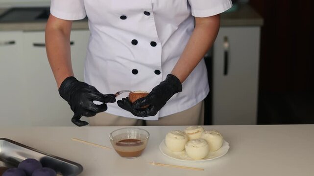 Female pastry chef dipping cake pop into chocolate glaze in modern kitchen