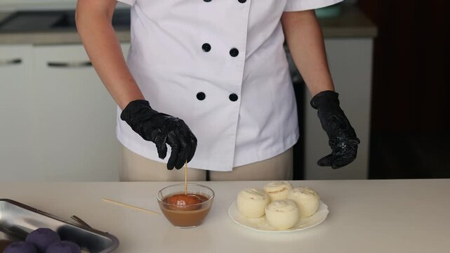 Female pastry chef dipping cake pop into chocolate glaze in modern kitchen