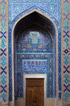 Ornate tiled portal with pointed arch and carved wooden door at Shah-i-Zinda necropolis in Samarkand. Intricate Islamic tilework featuring geometric and floral patterns in blue and turquoise.